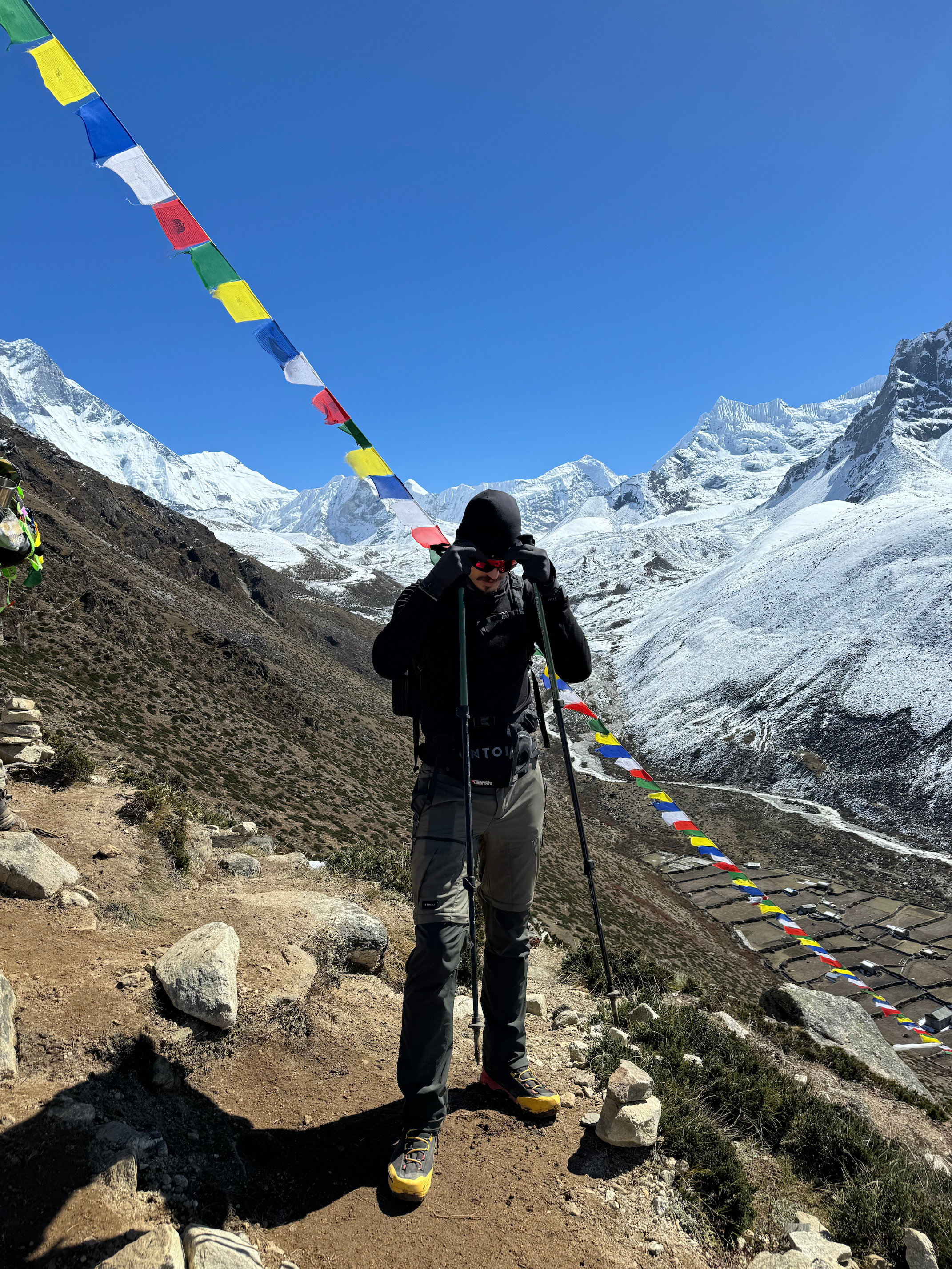 Hiker standing on a rocky trail beneath snow-covered Himalayan peaks and colorful prayer flags in Nepal